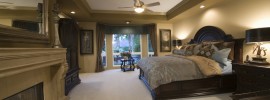 This beautiful luxurious rich golden colored room has dark wood furnishing, classic fireplace, and neutral bed furnishings